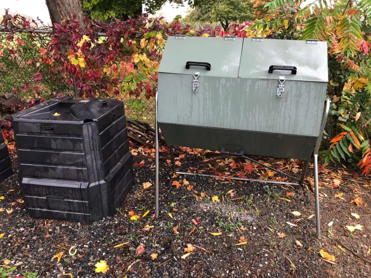 A metal round bin on legs and a plastic cube bin stand side by side in front of a fence with vines growing along it that are turning fall colors of crimson and yellow. 