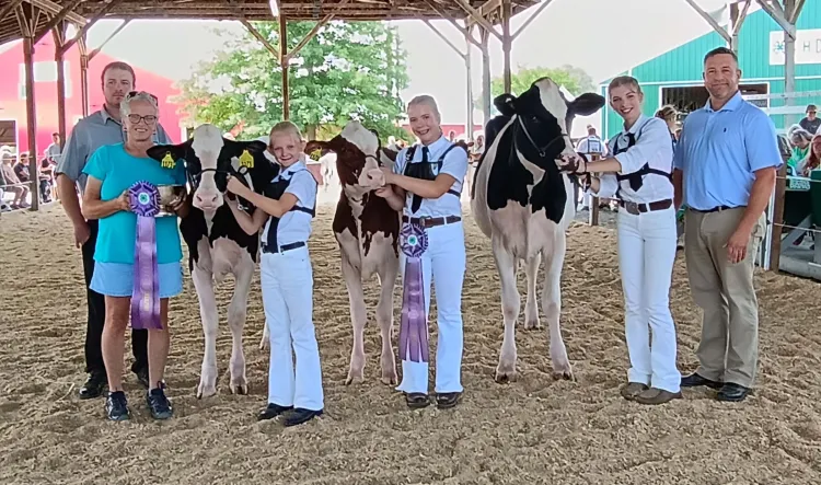 Six people and three cows stand for a portrait inside a show ring with a roof. The three middle people are younger teen and preteen girls who are holding on to cows by their halter. 