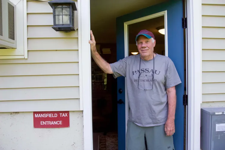 A white man in a baseball cap and t-shirt stands in a doorway of a home next to a red sign reading Mansfield Tax Entrance. 