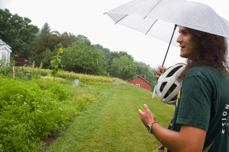 A person with reddish-light-brown skin long brown hair in a green t-shirt holds up an umbrella and a white bike helmet in a lush green garden under white-grey rainclouds.