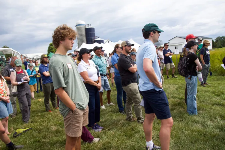 A group of people stand facing to the right while listening to someone speak outside on a grassy area in front of barn silos.