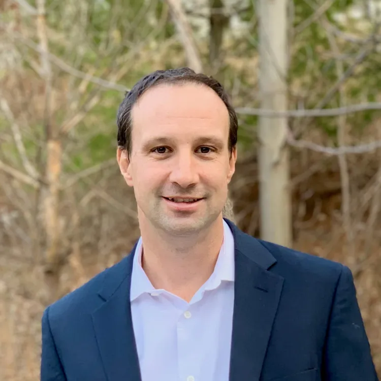 A headshot of a white man in a navy blue suit jacket and lavender button-down. He's standing outside with trees in the background.