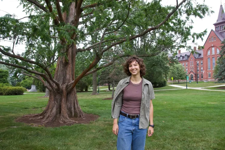 A young woman with light skin and red-brown shoulder-length curly hair stands in front of a tree on a grassy lawn in front of a large brick building. 