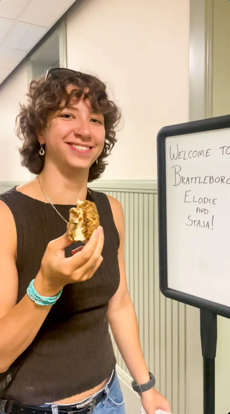 A young woman with light skin and red-brown shoulder-length curly hair holds an ice cream sandwich and smiles while standing next to a white board that reads "Welcome to Brattleboro Elodie and Staja!"