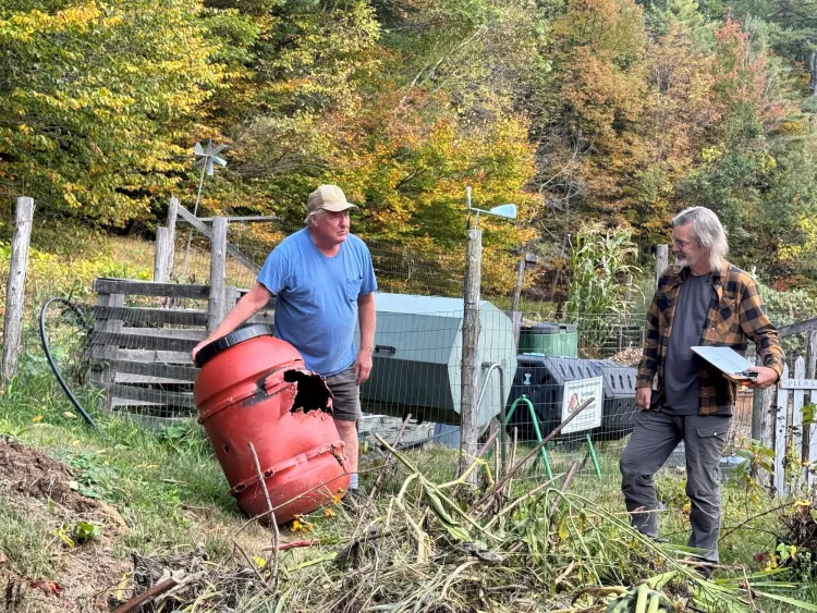 A light-skinned man in a blue t-shirt and yellow hat holds up a red barrel while a second light-skinned man in a flannel shirt looks on. They're both in a garden in early autumn.