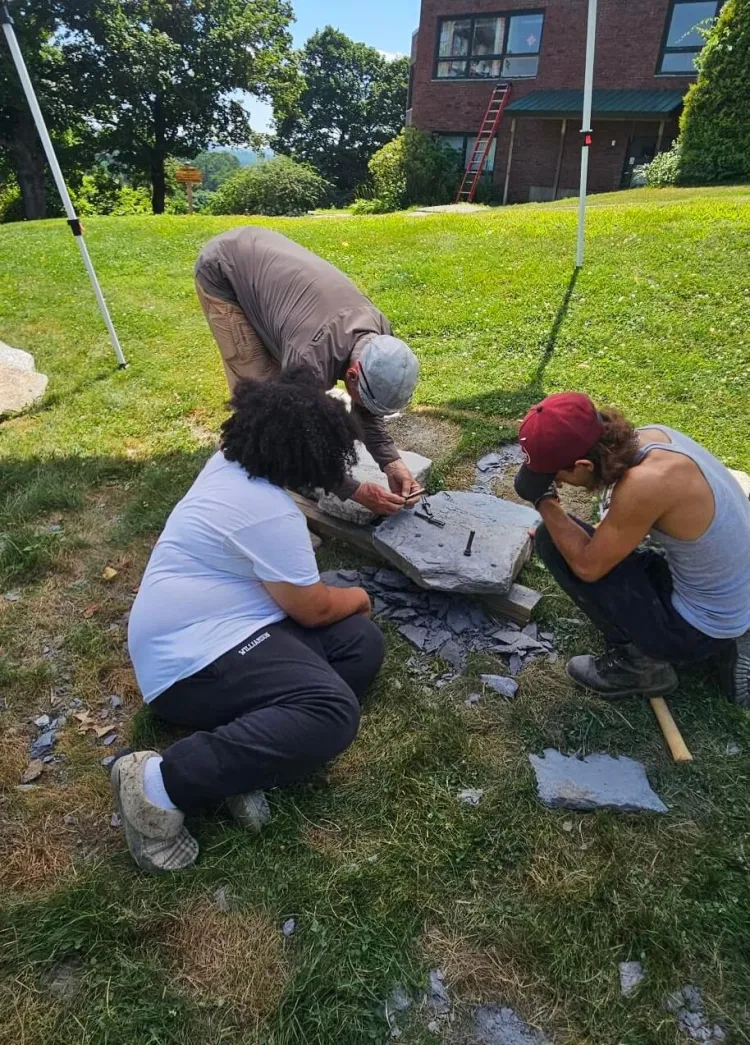 Three people sit and bend over around flat stones on the grassy ground in the shade of a tent. 
