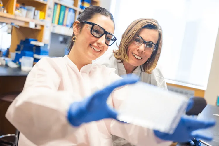 Two people in a laboratory setting in white lab coats and blue rubber gloves looking and smiling at a tray that the person on the left is holding up. 