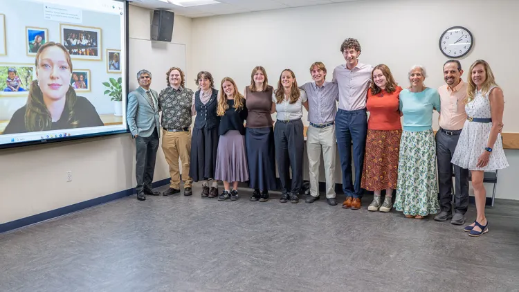 Students and faculty gather in a classroom for a group photo with one student appearing on a projection screen.