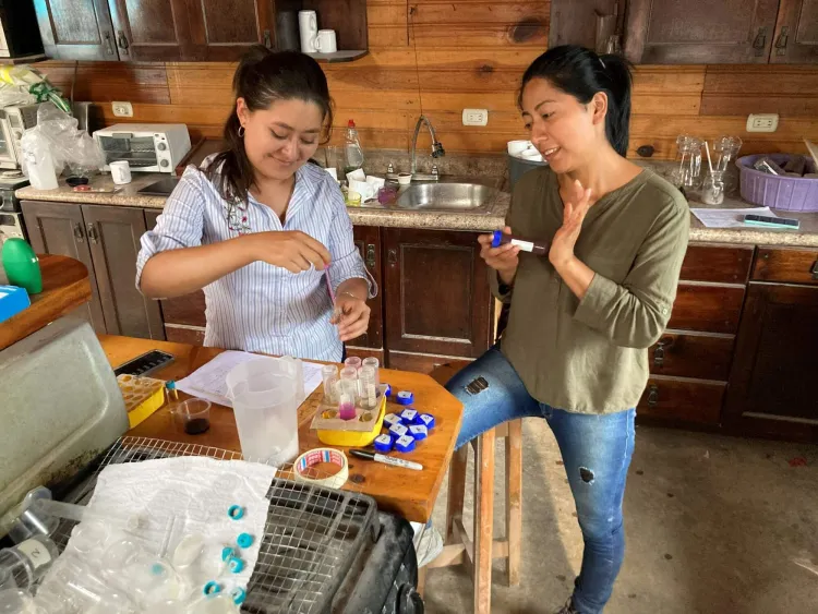 Two women holding test tubes stand around a table.