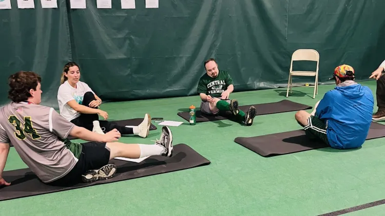 Participants sit on exercise mats in an indoor gym while taking part in a guided stretching session.  Caption:  Athletes gather on mats for a stretching and mobility activity as part of a Special Olympics fitness program.