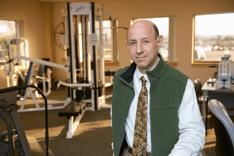 A person in a green vest and formal attire stands and smiles at the camera in a workout facility. 
