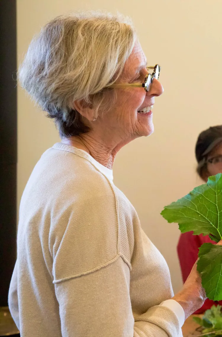 A white woman with short blonde hair and glasses smiles while holding a plant.