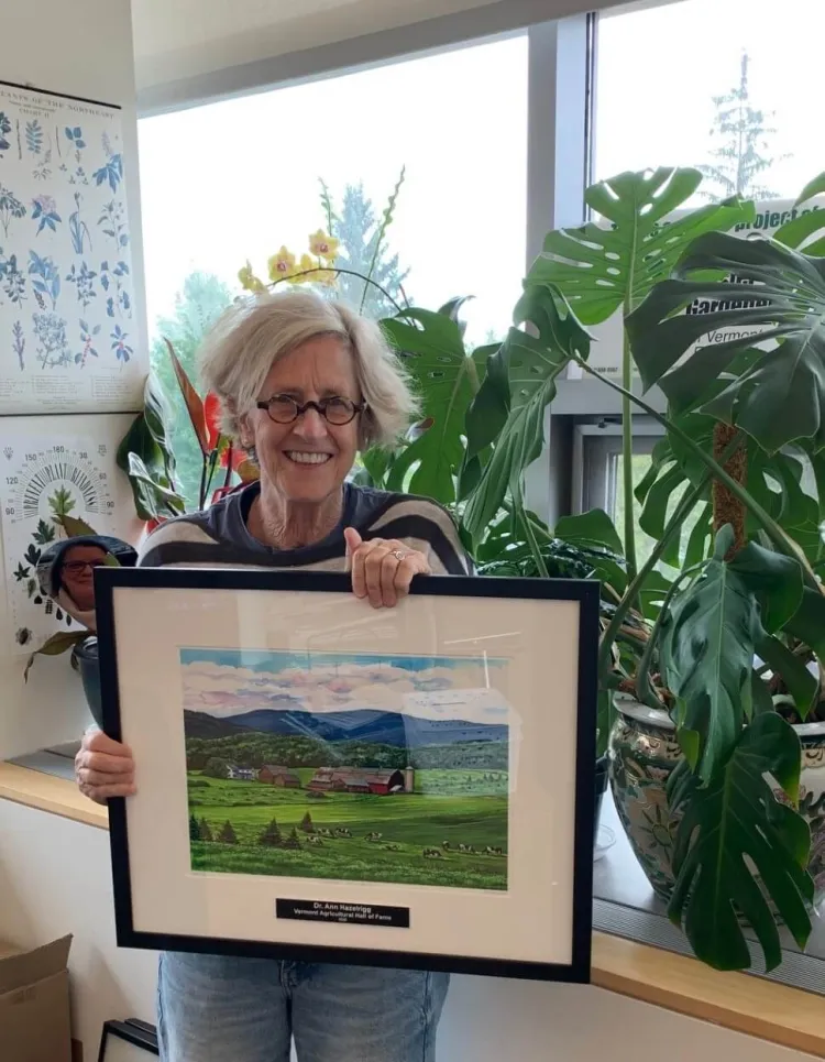 A white woman with short blonde hair smiling and holding a large framed painting of green fields and mountains.