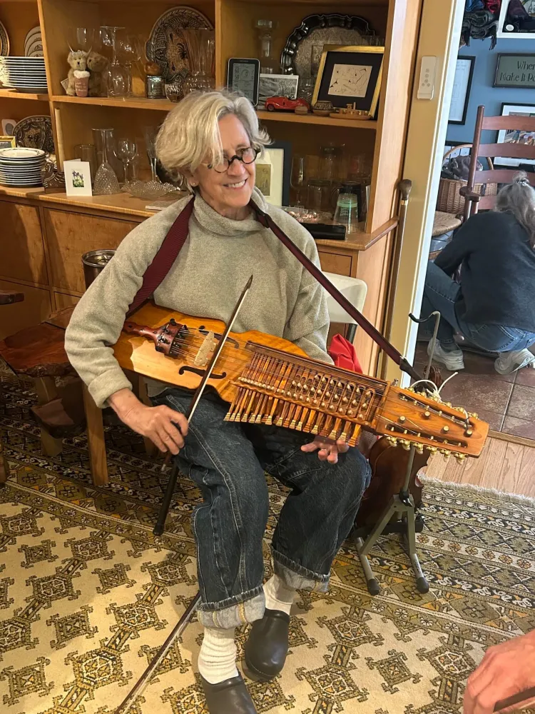 A blonde woman in glasses in a turtleneck holding a stringed, violin-like instrument that also has a neck with keys on it.