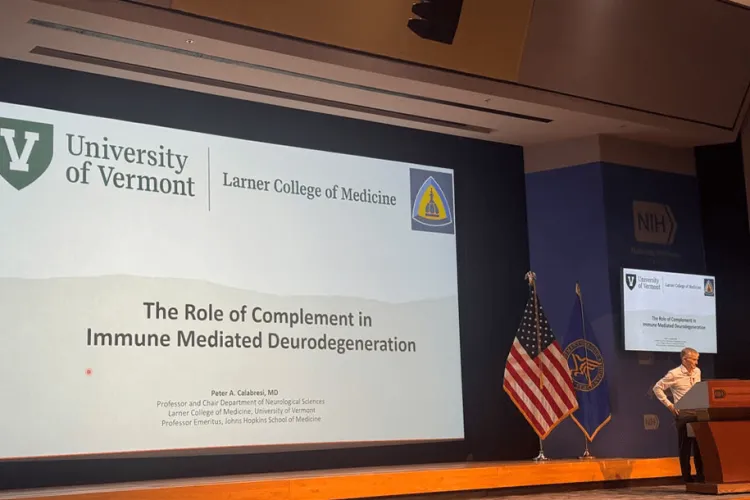 Large screen with presentation visible and a person at the podium. The presentation slide reads "The Role of Complement in Immune Meditated Neurodegeneration."