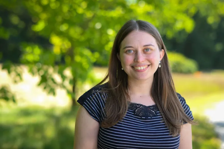 A light-skinned woman with long brown hair and a striped t-shirt smiles outside in front of sunlit greenery. 