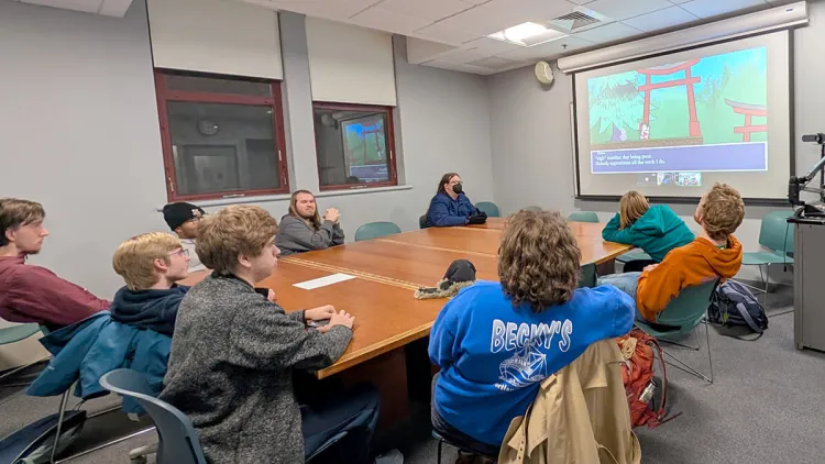 a group of students gathered around a conference table with an image of a video game projected on a large screen