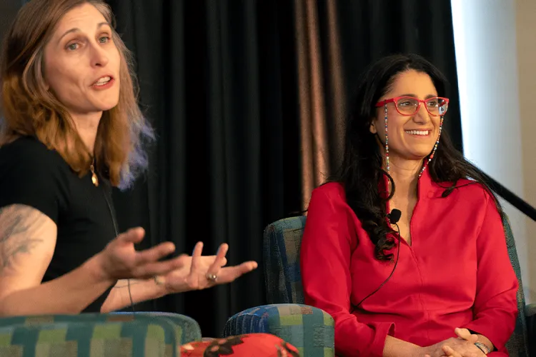 Two individuals sitting in chairs not facing the camera but turned to an audience. The one on the right is wearing a bright red shirt and the one on the left is in black. 