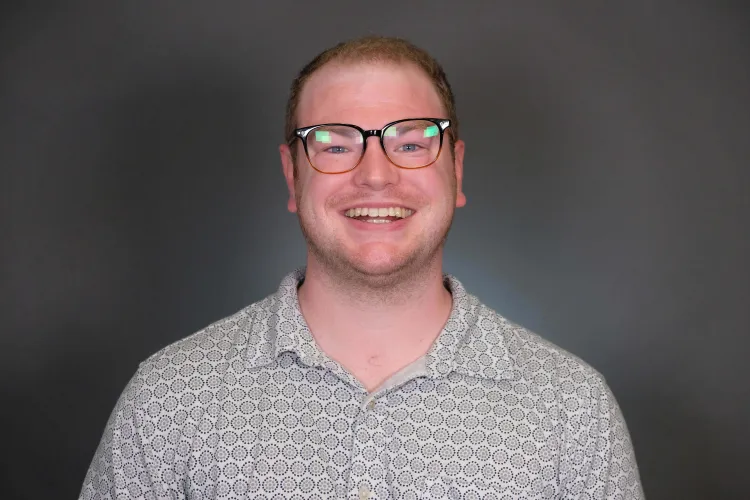 Person in a grey polo shirt and in glasses smiling at the camera with a grey background.