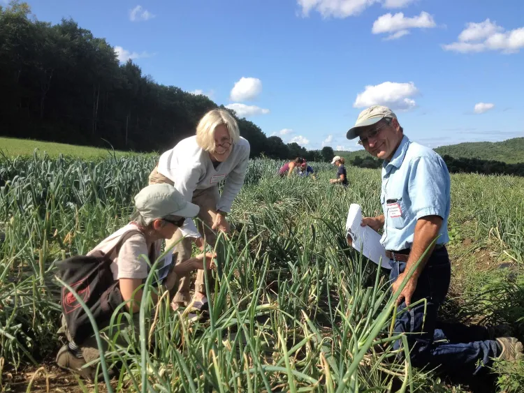 People in a long field with crops growing, and a blonde woman with glasses smiling into the camera.
