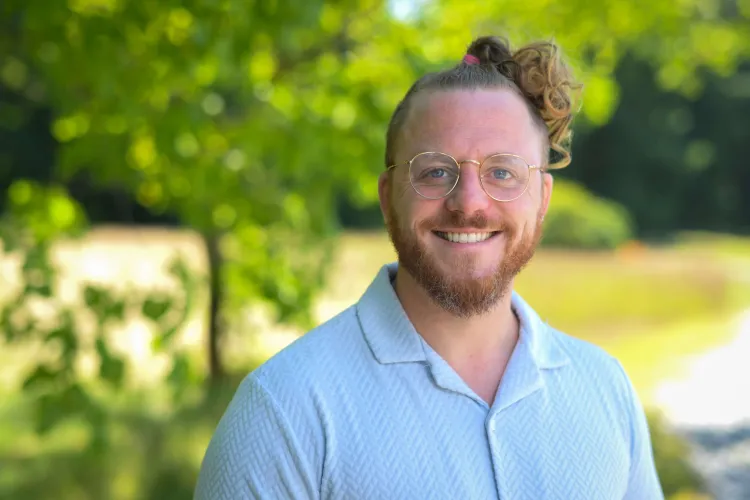 A light-skinned person with a strawberry blond beard and topknot hairstyle and glasses, in a light blue shirt against sunny greenery in the background.