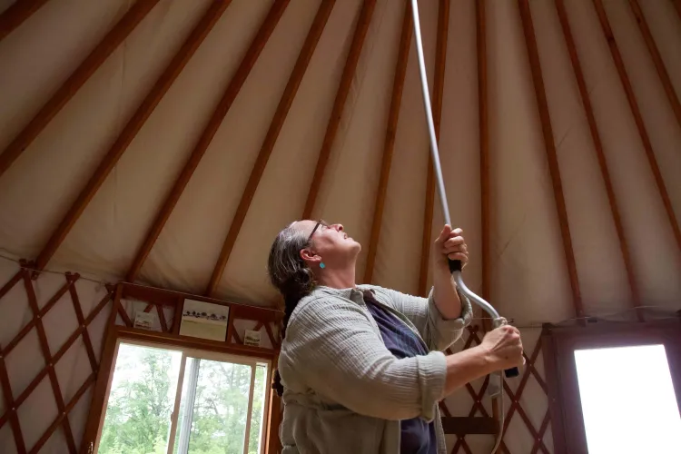 A woman with light skin and gray braided hair and glasses holds a rod roof-ward inside a yurt, which is held up by a lot of narrow strips of wood. 