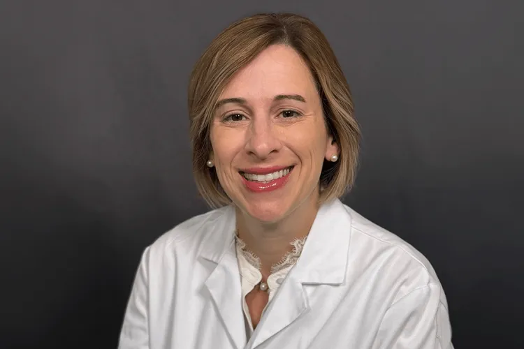 Person in a white lab coat against a grey background smiling at the camera for a headshot. 
