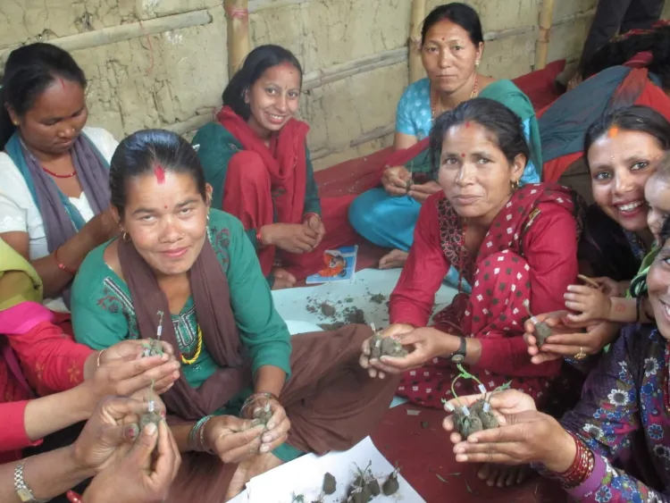 A group of women with brown skin and black, pulled-back hair and colorful dresses hold up plants in their hands and smile.