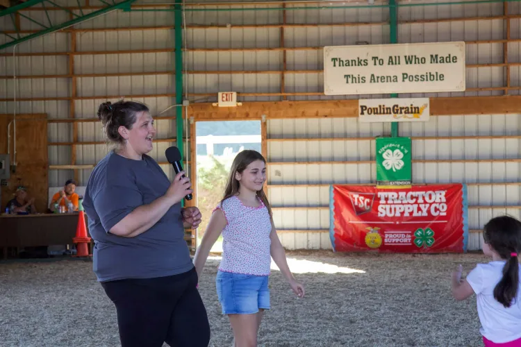 A light skinned woman in a grey shirt and black pants holds a microphone and speaks next to a girl with light skin in a light pink shirt and denim shorts. They're inside a show barn.