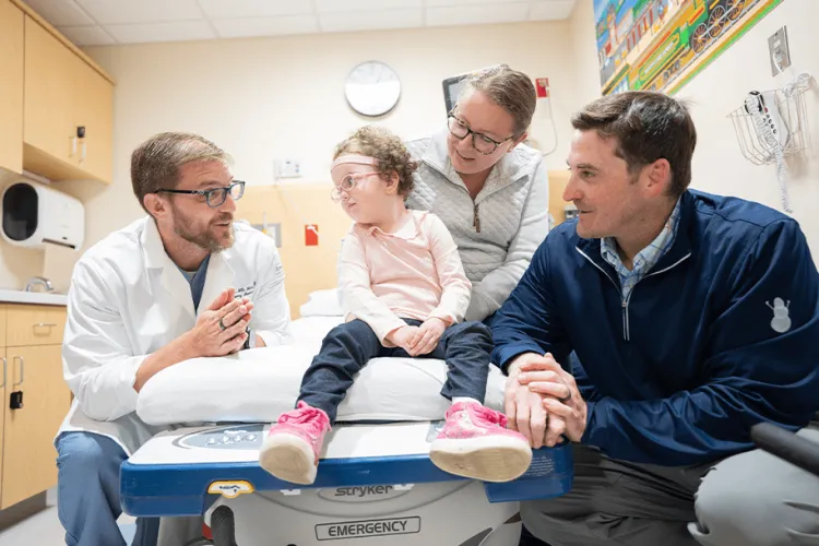 Doctor in a lab coat kneeling down to a child sitting on a hospital bench with their parents to the right of them. The setting is a hospital room. 