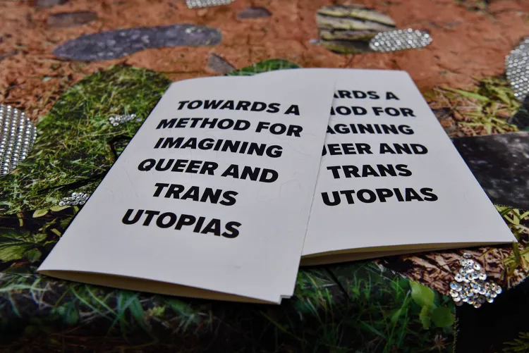Overlapping pamphlets on a multicolor background read, "Towards a method for imagining queer and trans utopias." Artwork by Jaimes Mayhew.
