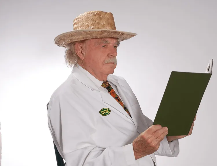 A man seated in a white lab coat with a straw hat reading a book