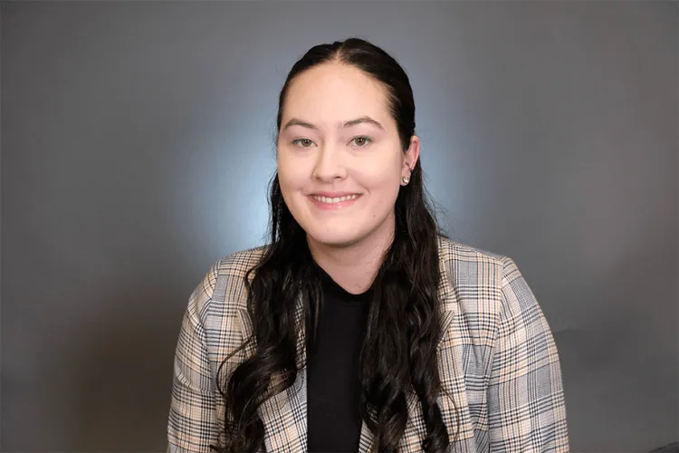 Person in a cream and navy plaid blazer and long brown hair pulled back smiles at the camera with a grey background. 