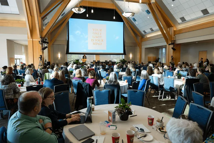 People in a conference room sitting around tables looking at a screen. 