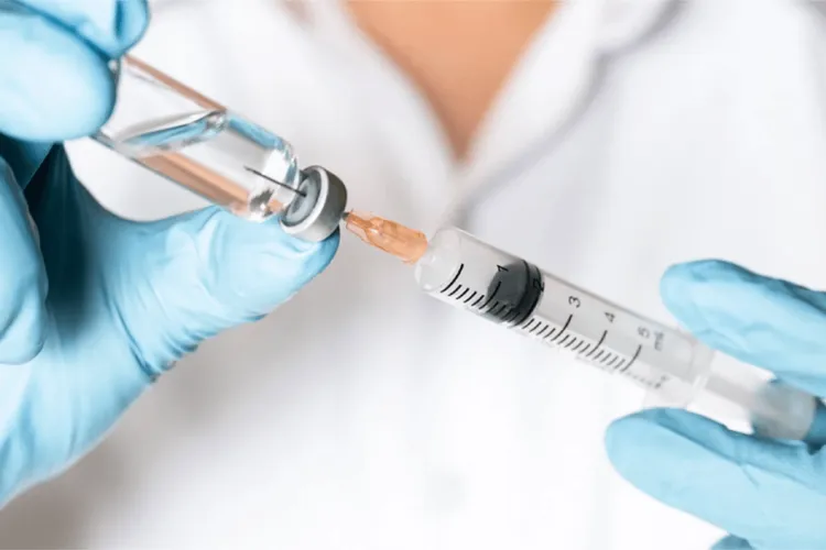 Close-up on a person in a lab coat with blue sterile gloves filling a syringe from a small glass vial. 