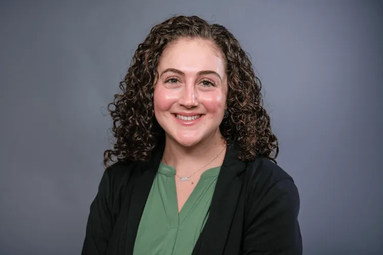 A person in a green shirt and black blazer smiles at the camera against a grey background in a photo studio. 