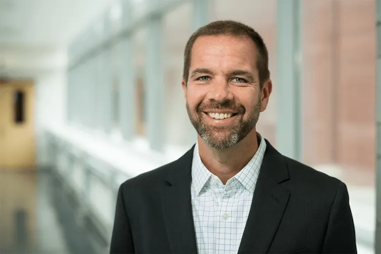 A person smiling at the camera in a glass hallways in a blue button up shirt with a black blazer. 