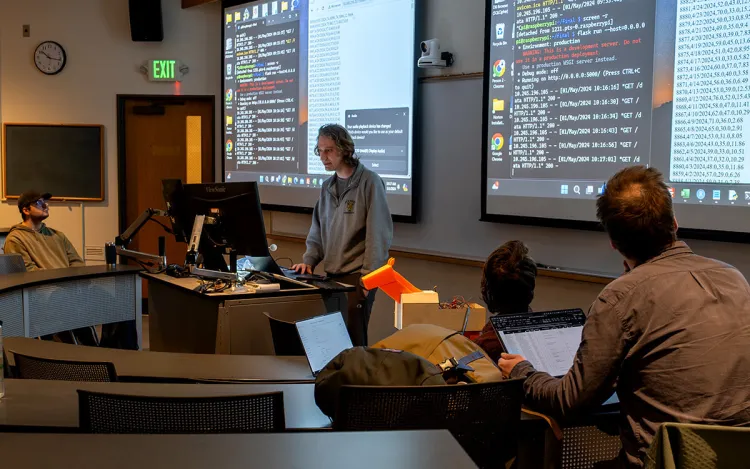A student presenting at a lectern in a classroom with a projection of computer code on screens behind him