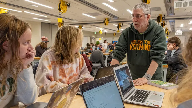 A professor speaks with several students around a table with open laptops