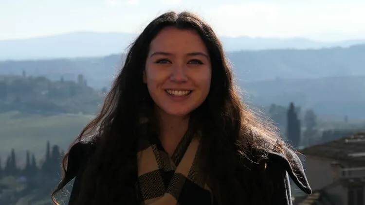 Smiling person with dark hair standing outdoors with hills in the background.