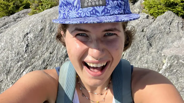 Emma Campbell smiling outdoors while wearing a purple floral-patterned hat and a backpack, with large rocks in the background.