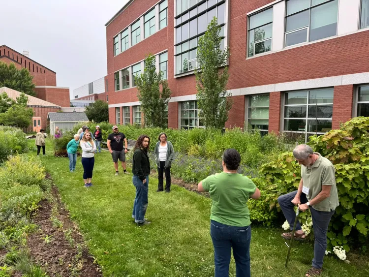 About a dozen people stand on a strip of grass next to a brick building.