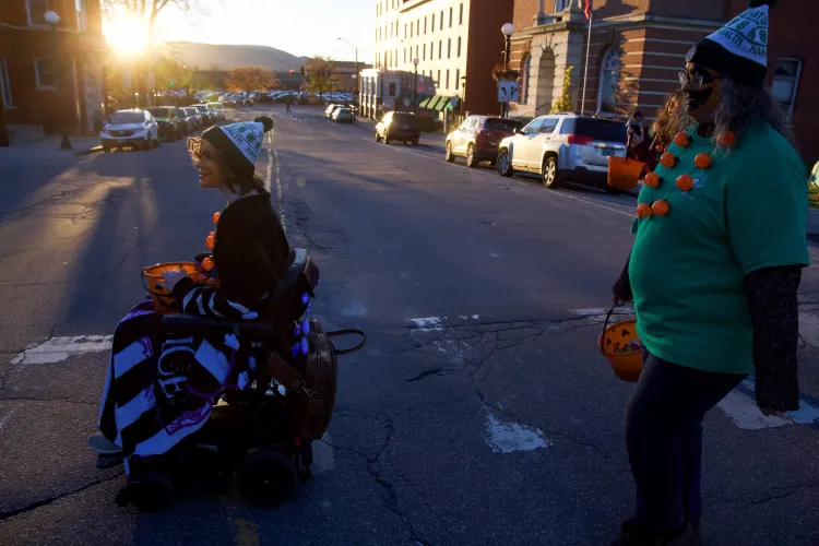 A photo of a young person and an adult, one using an electric wheelchair, the other walking, and both wearing white and green 4-H hats, cross a crosswalk on a downtown street during sunset.