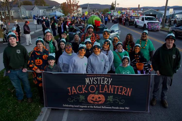 A group of people of all ages stand in front of a banner reading "mystery jack o'lantern" and wearing green and white hats.