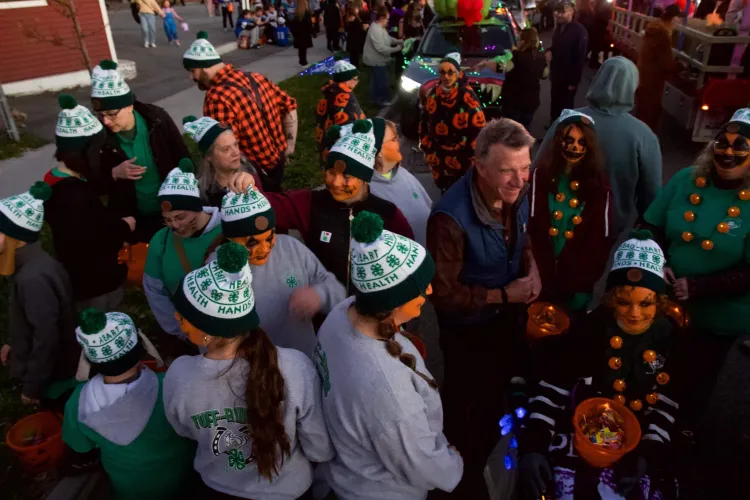 A man in a vest and flannel shirt stands among children and adults in 4-H green and white hats, with orange facepaint.