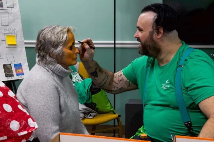 A white man in a green shirt and suspenders paints the face of an older white woman orange, like a pumpkin.