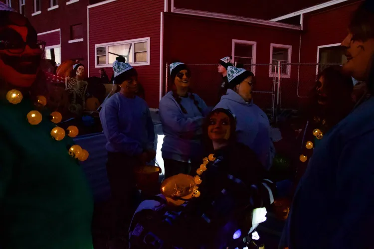 People in white and green 4-H hats, orange face paint and glowing pumpkin necklaces gather outside a red building.