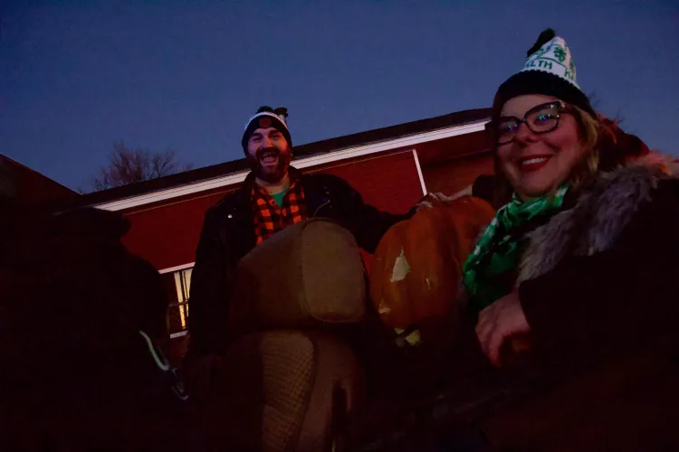 Two people smile in the backseat of an open-topped car, against a purplish-blue dusky sky. 