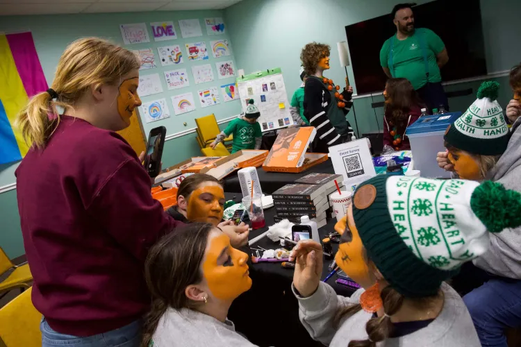 Youth and a couple adults paint their faces orange inside a green-walled room.  