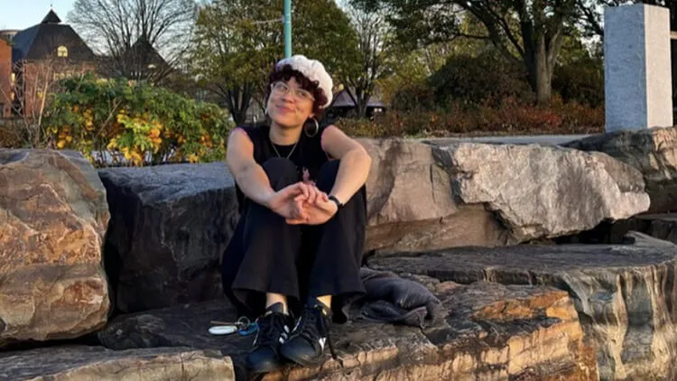 A person wearing a white hat and black outfit sits smiling on rocks outdoors.
